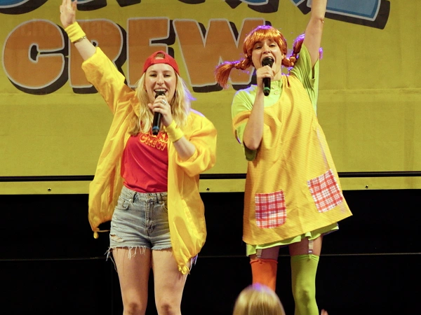 Zwei Sängerinnen der DONIKKL Crew singen mit Mikrofonen und erhobenen Armen bei einem Kinderkonzert.